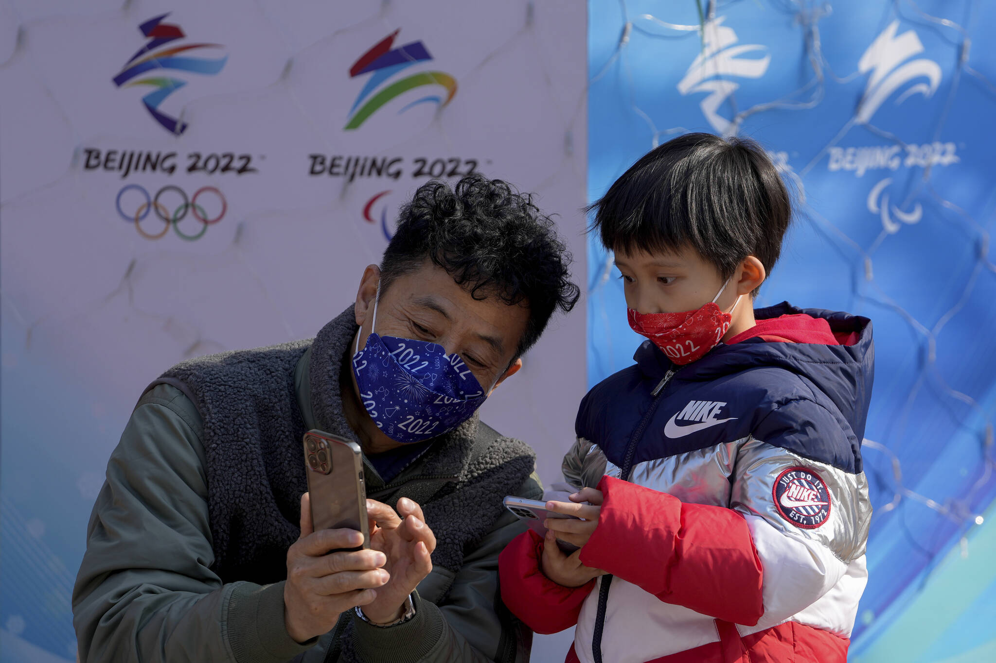 A man wearing a face mask to help protect from the coronavirus teaches a masked child how to take smartphone picture near the Beijing Winter Olympics decorations at the Wangfujing pedestrian mall in Beijing, Wednesday, Feb. 9, 2022. (AP Photo/Andy Wong)