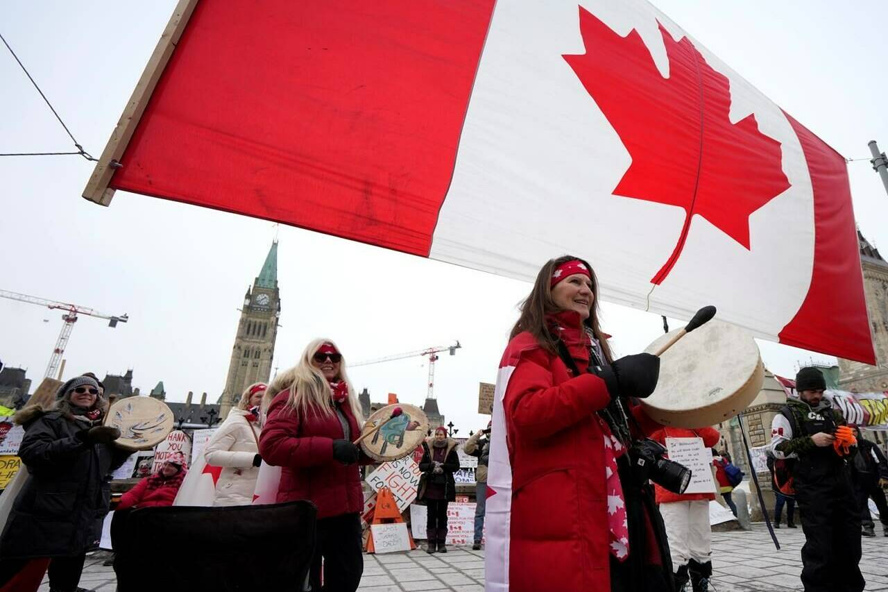 Women drum as they pass under a giant Canadian flag in front of Parliament Hill on the 20th day of a protest against COVID-19 measures that has grown into a broader anti-government protest, in Ottawa, on Wednesday, Feb. 16, 2022. Lawyers for a proposed class-action against the convoy protest in Ottawa said in an Ontario court today that GiveSendGo may be in breach of a freezing order on funds in connection with the convoy's fundraising efforts.THE CANADIAN PRESS/Justin Tang
