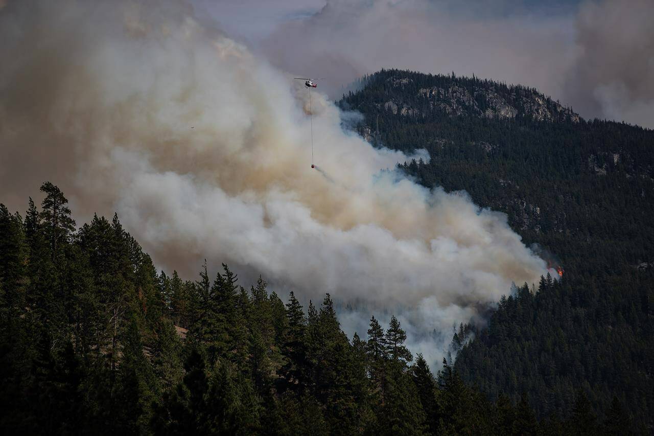 A helicopter carrying a water bucket flies past the Lytton Creek wildfire burning in the mountains near Lytton, B.C., on Sunday, August 15, 2021. A report on the wildfire that destroyed the British Columbia village of Lytton concludes the disaster couldn’t have been stopped, even with an area-wide emergency response.THE CANADIAN PRESS/Darryl Dyck