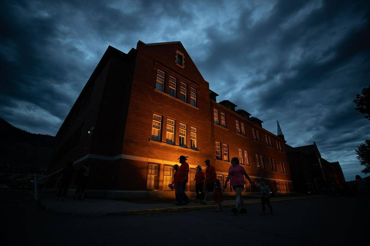 People are silhouetted as they walk past the former Kamloops Indian Residential School after gathering to honour the 215 children whose remains have been discovered buried near the facility, in Kamloops, B.C., on Monday, May 31, 2021. The year since the the Tk’emlups te Secwepemc First Nation announced that ground-penetrating radar had located the suspected grave sites in a former apple orchard has been one of national reckoning about residential schools in Canada. THE CANADIAN PRESS/Darryl Dyck