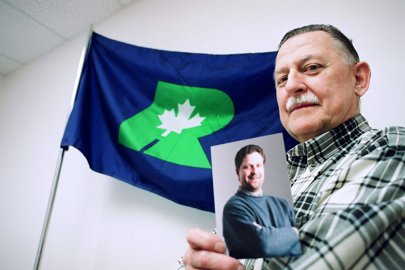 Duncan Goguillot is seen holding a photo of Chuck Strahl on Jan. 31, 2000 in front of a Reform party flag. (Jenna Hauck/ Chilliwack Progress file)