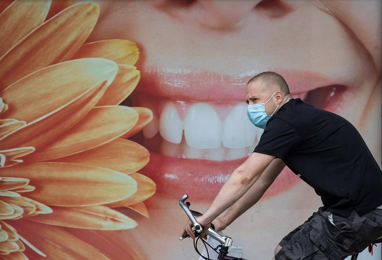 A man wearing a face mask to curb the spread of COVID-19 rides a bike past a photograph of a woman smiling outside a dental office, in Vancouver, B.C., Monday, Aug. 3, 2020. Economic and public policy experts warn the federal dental benefit may not reach the families who are most in need because the government has chosen to distribute it through the tax system. THE CANADIAN PRESS/Darryl Dyck