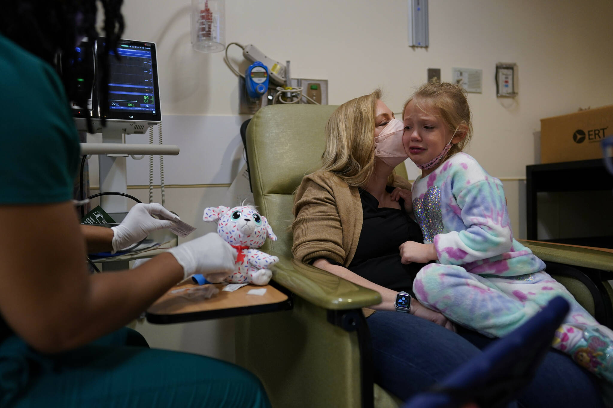 Kate Forte kisses her daughter, Lexie Stroiney, 6, as research nurse Michelle Harris, left, demonstrates a blood draw on her stuffed animal “Sprinkles” at Children’s National Hospital, in Washington, Wednesday, Jan. 26, 2022. Lexie had COVID-19 and is part of a NIH-funded multi-year study at Children’s National Hospital to look at impacts of COVID-19 on children’s physical health and quality of life. (AP Photo/Carolyn Kaster)