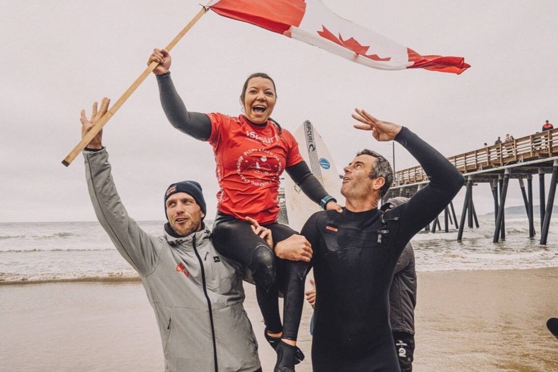 Victoria Feige is carried out of the surf and onto the podium by Team Canada coaches Albie Sears, left, and Shannon Brown after winning her fourth women’s kneel world title on Dec. 10. (ISA Pablo Franco photo)
