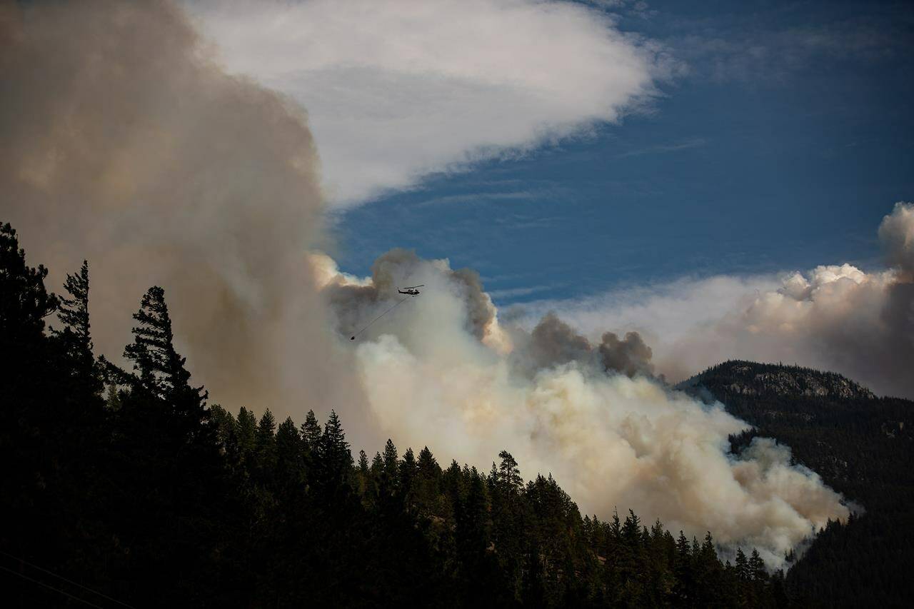 A helicopter carrying a water bucket flies past the Lytton Creek wildfire burning in the mountains near Lytton, B.C., on Sunday, Aug. 15, 2021. Two out-of-control wildfires in northeastern British Columbia that have already forced some residents to evacuate their homes are expected to grow bigger in the next few days. THE CANADIAN PRESS/Darryl Dyck