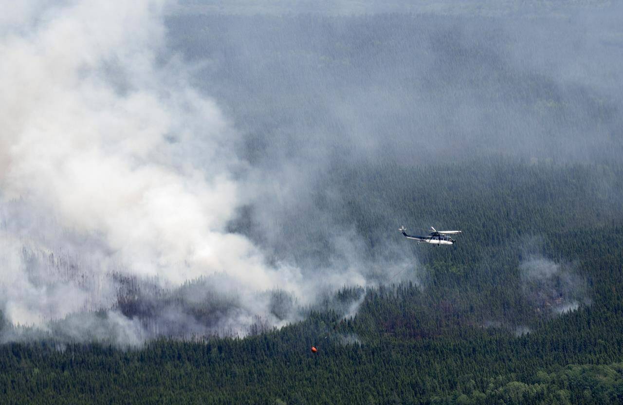 A helicopter carrying a water basket flies past a smoke plume near Lebel-sur-Quevillon, Que., Wednesday, July 5, 2023. Natural Resources Canada says the country’s record-breaking wildfire season will continue to be greater than normal throughout July and into August. THE CANADIAN PRESS/Adrian Wyld