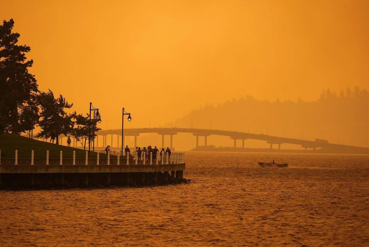 A person travels in a boat past people walking on the boardwalk as smoke from the McDougall Creek wildfire blankets the area on Okanagan Lake, in Kelowna Friday, Aug. 18, 2023. (THE CANADIAN PRESS/Darryl Dyck)
