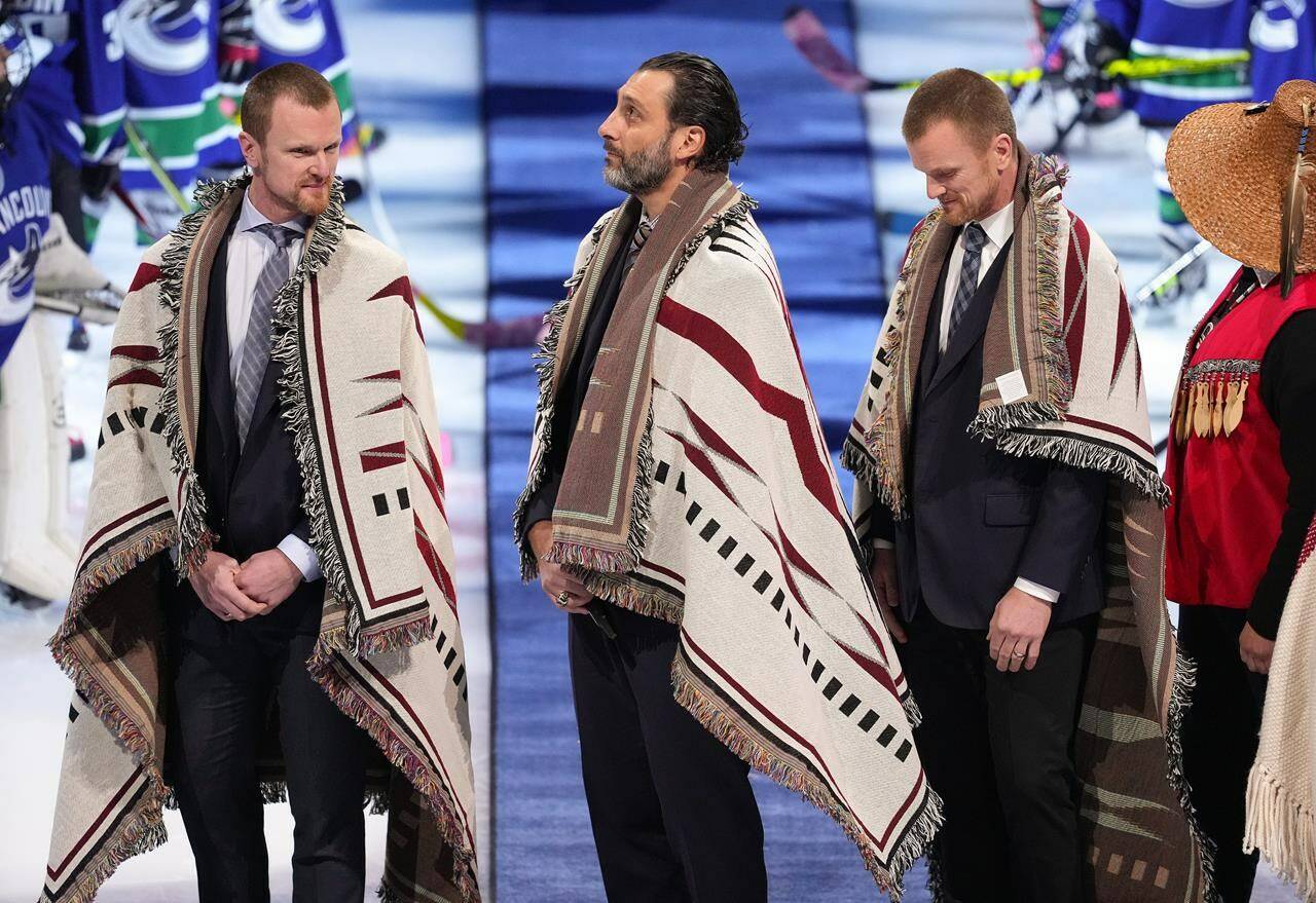 Former Vancouver Canucks Henrik Sedin, left, of Sweden, goalie Roberto Luongo, centre, and Daniel Sedin, right, of Sweden, stand after being wrapped in First Nations blankets during a ceremony honouring their recent inductions into the Hockey Hall of Fame, before an NHL hockey game in Vancouver, on Thursday, December 1, 2022. THE CANADIAN PRESS/Darryl Dyck