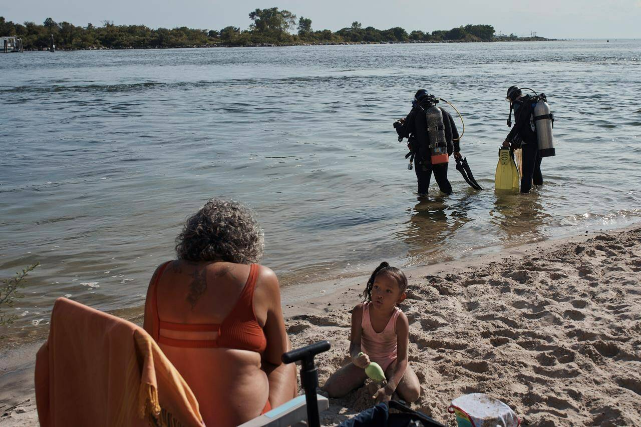 A family spends time on the beach as scuba divers, Tanasia Swift, second right, and Sarah Sears first right, prepares to enter the water during an underwater cleanup in the Queens borough of New York on Sunday, Aug. 27, 2023. A diving group takes part in a monthly cleanup at a cove in the community of Far Rockaway, about 4 miles south of John F. Kennedy Airport, to help the global effort to undo ocean pollution. (AP Photo/Andres Kudacki)