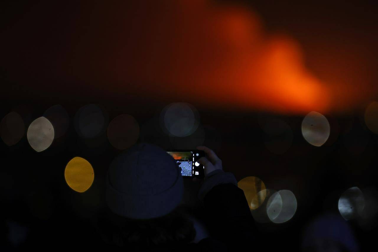 People take images as the night sky is illuminated caused by the eruption of a volcano on the Reykjanes peninsula of south-west Iceland seen from the capital city of Reykjavik, Monday Dec. 18, 2023. (AP Photo/Brynjar Gunnarsson)