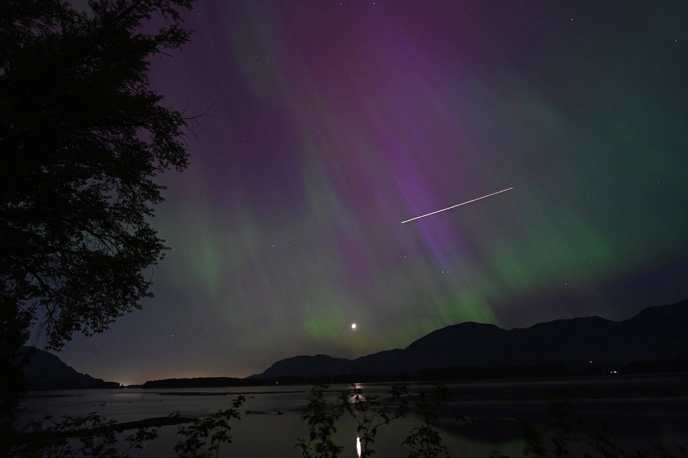 An airplane cuts through the sky as the northern lights are seen over the Fraser River at Old Orchard Road in Chilliwack on Friday, May 10, 2024. (Jenna Hauck/ Chilliwack Progress)