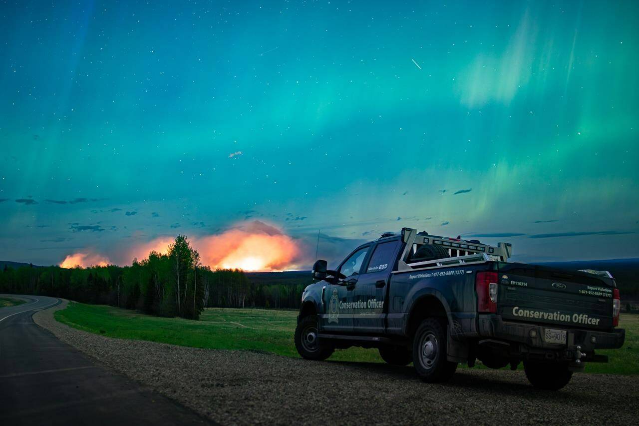 The mayor of a northeast British Columbia municipality where thousands have already evacuated says his community is bracing for a “last stand” as high winds are expected to push a nearby wildfire directly toward the town. The Aurora Borealis shines overhead of a B.C. Conservation Officer Service vehicle near the junction of highways 97 and 77, as a wildfire burns in the background near Fort Nelson, B.C., in a Saturday, May 11, 2024, handout photo. THE CANADIAN PRESS/HO-Ministry of Water, Land and Resource Stewardship