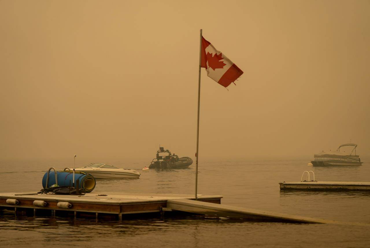 Thick smoke from the Lower East Adams Lake wildfire fills the air and a Canadian flag flies in the wind as RCMP officers on a boat patrol Shuswap Lake, in Scotch Creek, B.C., on Sunday, August 20, 2023. THE CANADIAN PRESS/Darryl Dyck